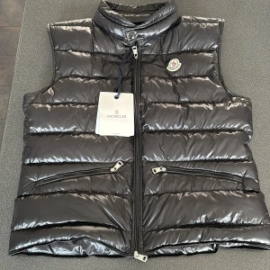 Moncler gui väst - Snygg svart dunväst från Moncler med glansig finish och klassisk logga på bröstet. Västen har hög krage, två snedställda fickor med dragkedja och quiltad design. Perfekt för lager-på-lager och streetstyle. Storlek 2 som motsvarar M