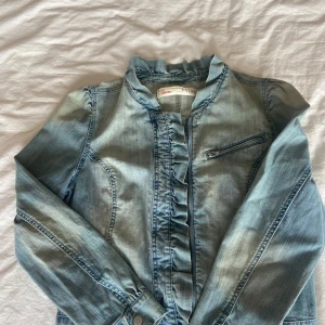 Ljusblå jeansjacka  - Snygg ljusblå jeansjacka med volangdetaljer vid krage och nederkant. Jackan har lång ärm, markerade sömmar och en bröstficka med dragkedja. Perfekt för dig som vill ha en cool men söt jeansjacka. 