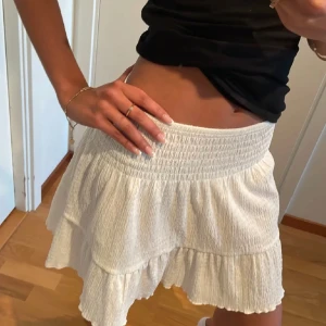 Vit volangkjol med resårmidja - Supersöt vit kortkjol med volanger och smockad resår i midjan. Kjolen har en luftig känsla och är perfekt för varma dagar. Den är tillverkad i ett lätt, strukturerat tyg som ger en avslappnad och trendig look.