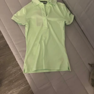 Ljusgrön golftröja från J.Lindeberg - Snygg ljusgrön golftröja från J.Lindeberg med klassisk krage och knappar framtill. T-shirten har korta ärmar och är tillverkad i ett mjukt, stretchigt syntetmaterial som passar perfekt för en sportig look. Diskret logga på kragen och stilren design.