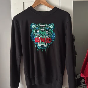 Svart Kenzo sweatshirt med tigerbrodyr - Svart sweatshirt från Kenzo med ikonisk tigerbrodyr i grönt, blått och rött framtill. Tröjan har rund halsringning, långa ärmar och ribbade muddar. Perfekt statement-plagg för dig som vill sticka ut med en cool och färgstark detalj.
