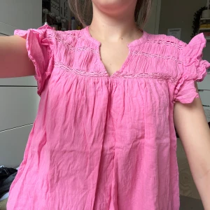 Rosa blus med volang från Gina Tricot - Supersöt rosa blus från Gina Tricot med volangiga korta ärmar och broderade detaljer upptill. Blusen är luftig och har en lätt rynkad struktur samt v-ringning. Perfekt för dig som gillar en feminin och chill stil.