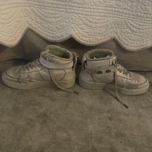 Vita höga sneakers från HOPE - Vita höga sneakers från HOPE med snörning och knappar upptill. Skorna har mjukt foder för extra värme och komfort. Rund tå och platt sula, perfekta för kyligare dagar.