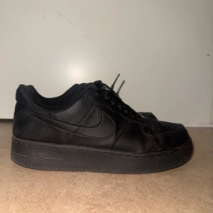 Svarta Nike Air Force 1 sneakers - Klassiska helsvarta Nike Air Force 1 sneakers i skinn med lågt skaft och perforerad tå. Snygg och clean look med svarta snören och metallplatta framtill. Perfekta för dig som gillar streetstyle och vill ha en tidlös sneaker i garderoben.