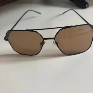 Svarta solglasögon med bruna glas - Snygga solglasögon med svart metallbåge och bruna, lätt tonade glas. De har en modern, kantig form och tunna skalmar. Näskuddarna är transparenta för extra komfort. Perfekta för att lyfta din stil under soliga dagar.Köparen står för frakt och varan köps i befintligt skick😊