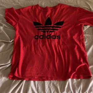 Röd Adidas t-shirt med svart logga - Röd t-shirt från Adidas med stor svart Trefoil-logga och text på bröstet. Klassisk passform med korta ärmar och rund hals. Tillverkad i mjuk bomull som är skön att ha på sig. Perfekt för en sportig och avslappnad stil.