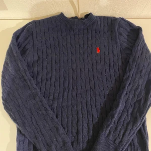 Mörkblå kabelstickad tröja Polo Ralph Lauren - Kabelstickad mörkblå tröja från Polo Ralph Lauren med klassisk röd broderad logga på bröstet. Tröjan har rund halsringning, långa ärmar och ribbade muddar. Perfekt för dig som gillar en tidlös och stilren look.