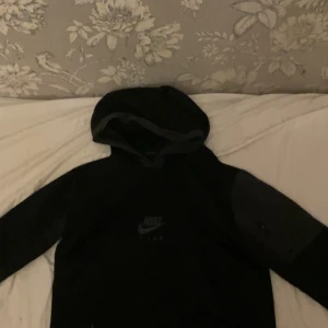 Svart Nike Air Max hoodie - Svart hoodie från Nike Air Max med diskret logga på bröstet. Tröjan har huva, långa ärmar och detaljer i mörkgrått på ärmarna. Perfekt för en sportig och avslappnad stil. Materialet känns mjukt och bekvämt, troligen polyester eller bomullsmix.