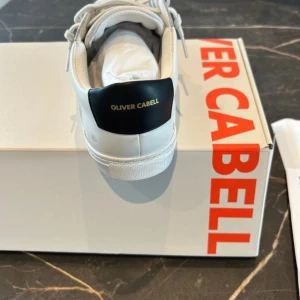 Vita sneakers från Oliver Cabell - Stilrena vita sneakers från Oliver Cabell med svart hälparti och guldfärgad logga. Skorna har ovandel i mocka och skinn, klassisk snörning och diskreta perforeringar på sidan. Perfekta för dig som gillar minimalistisk och clean stil.