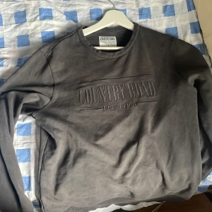 Mörkgrå sweatshirt från Country Road - Säljer en stilren mörkgrå sweatshirt från Country Road med broderad logga framtill. Tröjan har rund halsringning, långa ärmar och är tillverkad i mjuk bomull. Perfekt för en chill och avslappnad look.