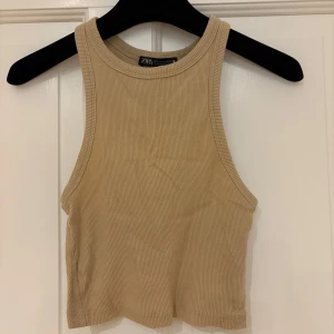 Beige ribbad linnetopp från Zara - Snygg croppad linnetopp från Zara i beige ribbat material. Toppen är ärmlös med hög halsringning och har en clean, stilren look som passar perfekt till jeans eller kjol. Superenkel att matcha och riktigt trendig för sommaren!