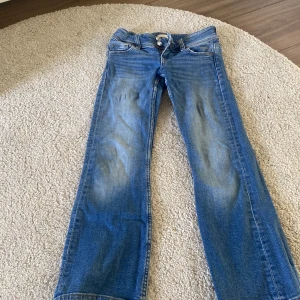 Blå bootcut jeans från Gina Tricot - Snygga blå jeans från Gina Tricot med bootcut-modell och klassisk femficksdesign. Jeansen har dubbla knappar i midjan och coola slitningar framtill. Tillverkade i jeansmaterial med kontrastsömmar och ficklock bak. 