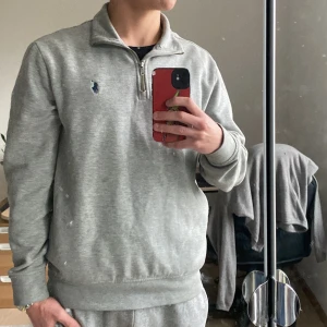 Grå halvzip sweatshirt från Polo Ralph Lauren - Säljer en stilren grå sweatshirt från Polo Ralph Lauren med halv dragkedja och broderad logga på bröstet. Tröjan har lång ärm och är tillverkad i mjuk bomullsmix. Perfekt för en avslappnad look. Passar även s. riktigt skön