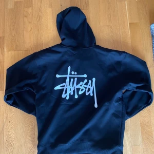 Svart hoodie från Stüssy - Säljer en svart hoodie från Stüssy med deras ikoniska logga tryckt i vitt på ryggen och framtill. Tröjan är otroligt skön eftersom att insidan är i silkes material. Perfekt för dig som gillar streetwear och vill ha något stilrent. Ej äkta