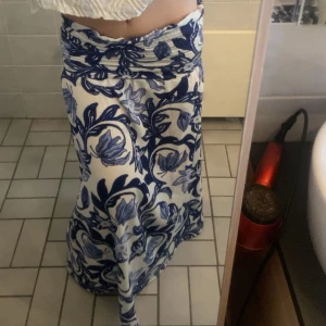 Vit och blå långkjol/ klänningmed blommigt mönster - Säljer en långkjol/klänning med vit bas och blått blommigt mönster. Kjolen har en bred linning och ett flowigt fall som ger en avslappnad känsla. Perfekt för varma dagar och enkel att styla med olika toppar. Passar s också. Oanvänd 