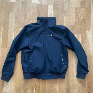 Marinblå vindjacka från Sail Racing - Säljer en marinblå vindjacka från Sail Racing med hög krage och dragkedja framtill. Jackan har två snedställda fickor med dragkedja och diskret logga på bröstet. Perfekt för blåsiga dagar.