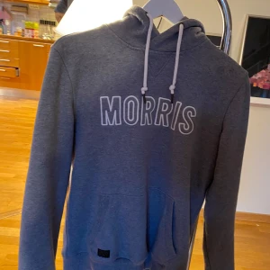 Grå hoodie från Morris - Säljer en grå hoodie från Morris med vit logga framtill och huva med dragsko. Tröjan har en klassisk känguruficka och ribbade muddar vid ärmslut och nederkant. Perfekt för en avslappnad stil.