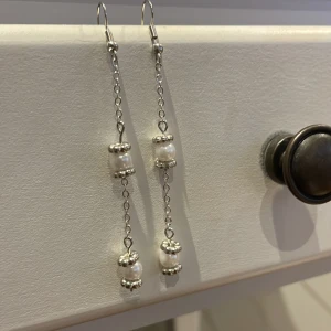 Långa silverörhängen med pärlor - Säljer ett par eleganta, långa örhängen i silverfärgad metall med kedja och vita pärlor. Örhängena har dekorativa detaljer runt pärlorna och ger en stilren och klassisk look.