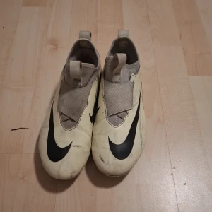 Nike Zoom Mercurial Vapor 15 Academy - Säljer ett par Nike Zoom Mercurial Vapor 15 Academy fotbollsskor i ljus gul med stora svarta swoosh-loggor och texten 'MERCURIAL' på hälen. Skorna har täckt snörning och plös, samt dobbar för spel på gräs. Perfekta för dig som vill sticka ut på planen.