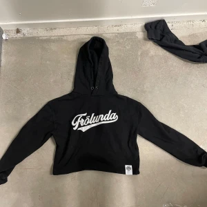 Svart hoodie med Frölunda-tryck - Svart hoodie med vit Frölunda-logga på bröstet. Tröjan har huva med dragsko och är croppad i modellen. Perfekt för dig som vill visa ditt stöd för Frölunda på ett stilrent sätt.