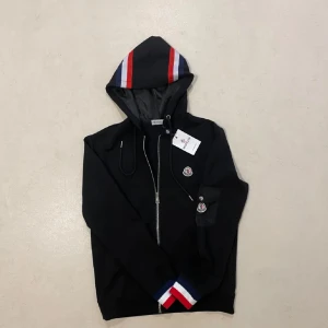 Svart hoodiejacka med dragkedja från Moncler - Svart hoodiejacka från Moncler med dragkedja framtill, huva och ribbade muddar i rött, vitt och blått. Jackan har Moncler-logga på bröstet och ärmen samt snörning i huvan. Perfekt för en sportig och stilren look.