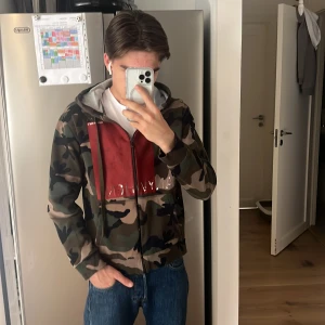 Camo hoodie från Valentino med röd detalj - Säljer en camouflagemönstrad hoodie från Valentino med röd fyrkant och text på bröstet. Tröjan har dragkedja, huva med snörning och känguruficka. Perfekt för dig som gillar streetwear och vill sticka ut.