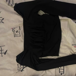 Svart croppad topp med rynkade detaljer - Säljer en svart croppad topp med långa ärmar och rynkade detaljer framtill. Toppen har en figurnära passform och är tillverkad i ett mjukt, stretchigt material. Perfekt att styla med höga jeans eller kjol.