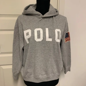 Grå hoodie från Polo Ralph Lauren - Säljer en grå hoodie från Polo Ralph Lauren med stor vit POLO-text på bröstet och amerikansk flagga på ärmen. Klassisk huva och ribbade muddar. Perfekt för en avslappnad stil. Storlek L i barn , passar mig S