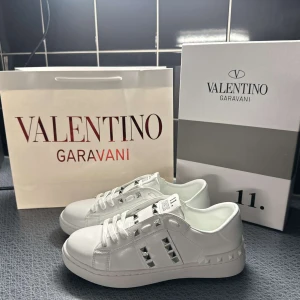 Vita Valentino Garavani Rockstud sneakers - Säljer ett par vita Valentino Garavani Rockstud sneakers med ikoniska nitar längs sidorna och på plösen. Skorna har snörning och är tillverkade i skinn med en platt sula. Perfekta för dig som vill ha en stilren men edgy look.