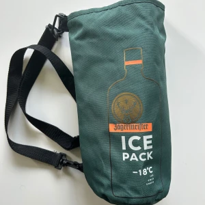 Grön Jägermeister axelväska med tryck - Cool grön axelväska från Jägermeister med flasktryck och texten 'ICE PACK -18°C' på framsidan. Väskan har justerbar svart axelrem och är fodrad med silverfärgat isolerande material på insidan. Perfekt för att hålla drycker kalla.