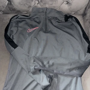 Grå träningströja med dragkedja från Nike - Säljer en grå långärmad träningströja från Nike med svart detalj längs ärmarna och halv dragkedja framtill. Tröjan har en röd Nike-logga på bröstet och är tillverkad i Dri-Fit material som andas.