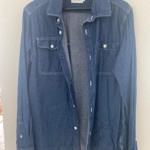 Jeansskjorta från East West - Säljer en klassisk blå jeansskjorta från East West med vita knappar och två bröstfickor. Skjortan har lång ärm och synliga sömmar som ger en snygg detalj. Perfekt att styla lager på lager.