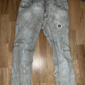 Grå jeansbyxor från Dsquared2 - Säljer ett par grå jeansbyxor från Dsquared2 med slitna detaljer, färgstänk och patch på ena benet. Byxorna har klassisk femficksmodell och knappgylf. Snygg streetstil med unika detaljer och Dsquared2-logga vid gylfen.