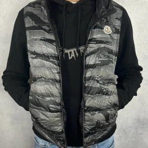 Moncler väst - Säljer en snygg dunväst från Moncler i svart och grått camouflage-mönster. Västen har huva, dragkedja framtill och två fickor med dragkedja. Moncler-logga på bröstet. Perfekt för lager-på-lager-stil.