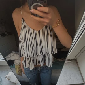 Randig volangtopp med tunna axelband - Säljer en luftig topp med svarta och vita ränder, volangdetaljer och tunna axelband. Perfekt för varma dagar och enkel att matcha med jeans eller kjol.