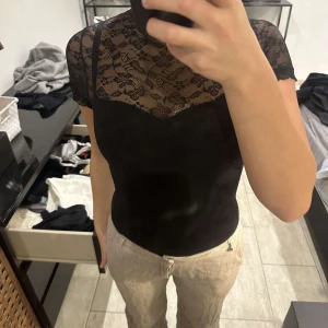 Svart spetsblus med hög hals - Säljer en svart blus med korta ärmar och hög hals i genombruten spets. Blusen har ett feminint och elegant mönster i spetsen och är figurnära i modellen. Perfekt att styla till både jeans och kjol.