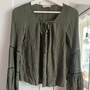 Olivgrön blus med vida ärmar och knytning - Säljer en olivgrön blus med långa, vida ärmar och dekorativa sömmar från Hollister. Blusen har v-ringning med knytning framtill och är tillverkad i ett lätt, skrynkligt tyg. Perfekt för dig som gillar boho-stil. Skriv för fler bilder💞