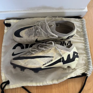 Nike Air Zoom Mercurial fotbollsskor - Säljer ett par Nike Air Zoom Mercurial fotbollsskor i beige och svart. Skorna har snörning, rund tå och platt sula med dobbar för bra grepp på planen. Tillverkade i syntetmaterial med ACC-teknologi och Vaporsposite+ för extra komfort och kontroll.