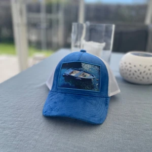 Velvet keps - Säljer en blå och vit keps med mesh-bak och justerbar snapback. Framsidan har ett tryck av en båt på vattnet. Perfekt för soliga dagar och en avslappnad stil. Kepsen är knappt använd och i nyskick.