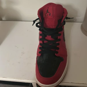 Nike Air Jordan 1 Mid röd och svart - Säljer ett par klassiska Nike Air Jordan 1 Mid i rött och svart med vita sulor. Skorna har svart snörning, svart swoosh och hög modell. Perfekta för dig som gillar streetwear och vill sticka ut med en ikonisk sneaker.
