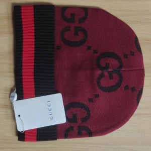 Röd mössa från Gucci med GG-mönster - Snygg röd mössa från Gucci med svart GG-logga runtom. Mössan har bred ribbad kant i mörkbrunt och rött. Perfekt accessoar för att sticka ut och hålla värmen.