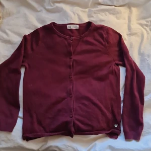 Vinröd kofta från H&M - Säljer en vinröd kofta från H&M i mjukt material. Koftan har rund halsringning, långa ärmar och knäppning framtill med små knappar. Perfekt att slänga över en topp för en enkel och stilren look.