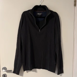 Mörkgrå stickad tröja med dragkedja från Indicode - Mörkgrå stickad tröja från Indicode med halv dragkedja och krage. Tröjan har långa ärmar och diskret broderad logga på bröstet. Perfekt för kyligare dagar och enkel att matcha med olika outfits. 