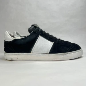 Valentino Garavani sneakers i svart och vitt - Snygga sneakers från Valentino Garavani i svart mocka med vita detaljer. Skorna har en rund tå och snörning framtill. Sulan har ett coolt kamouflagemönster och små nitar runt hälen för extra stilpoäng.