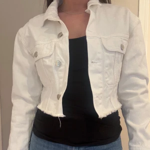 Vit croppad jeansjacka från NA-KD - Säljer en vit croppad jeansjacka från NA-KD med rå kant nertill och silvriga knappar. Jackan har klassisk krage, två bröstfickor och lång ärm. Endast testad, aldrig använt. Storlek 34, säljer för 100kr 🌸