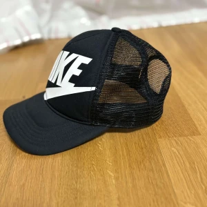 Nike keps - Svart keps från Nike med stor vit logga framtill. Kepsen har böjd skärm och baksida i mesh för extra ventilation. Justerbar passform med snapback-spänne. Perfekt för en sportig och avslappnad stil.
