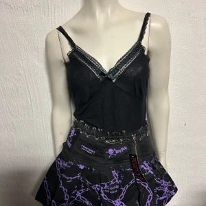 Svart och lila kjol med kedjemönster från Poizen Industries - Cool svart kjol med lila kedjemönster och spetskant nertill. Kjolen har dragkedja framtill, säkerhetsnålar som detalj i midjan och tryckt text samt dödskallemotiv. Märke: Poizen Industries.