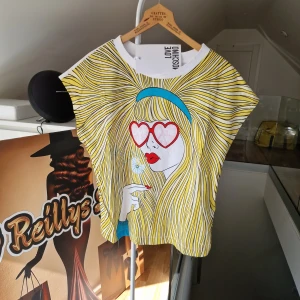 Färgglad t-shirt med pop art-tryck från Love Moschino - Cool t-shirt från Love Moschino med ett stort pop art-tryck av en tjej med blont hår, röda hjärtformade solglasögon och blått hårband. T-shirten har korta ärmar och är i en lös passform. Perfekt statement-plagg för dig som gillar färg och unika prints.
