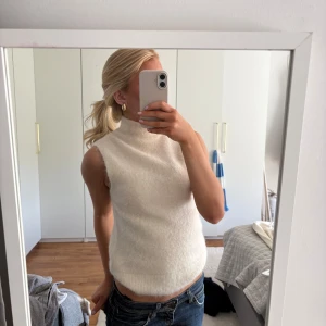 Vit ärmlös stickad polotopp - Säljer en vit, ärmlös stickad polotopp med hög hals. Toppen har en mjuk och mysig känsla och är perfekt att styla med jeans eller kjol. Enkel och stilren design som passar till många tillfällen.