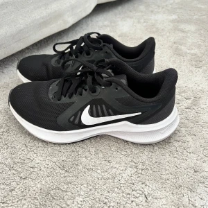Svarta Nike sneakers med vit sula - Säljer ett par svarta Nike sneakers med vit sula och vit swoosh-logga på sidan. Skorna har snörning och är tillverkade i syntetmaterial med mesh för bra andningsförmåga. Perfekta för träning eller vardagsstil.